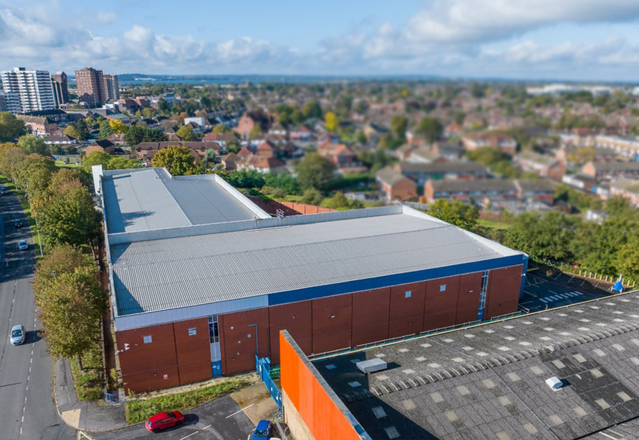 Aerial view of a large self storage facility with a blue roof in a residential area. A large parking lot filled with cars is in front of the building.