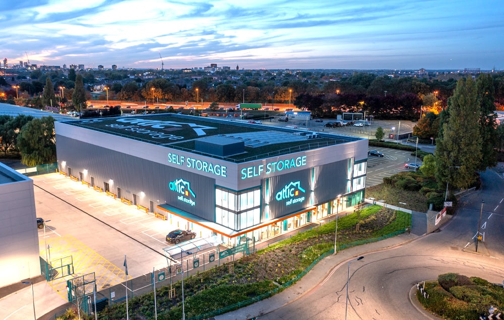 An aerial view of a self-storage facility at night. The text "Attic Self Storage" is written on the roof in teal letters.